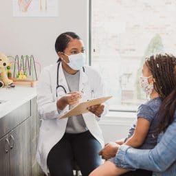 With a protective mask on, a female pediatrician talks to a young patient's mother about the woman's daughter's medical conditions. They are wearing protective masks during the COVID-19 pandemic.
