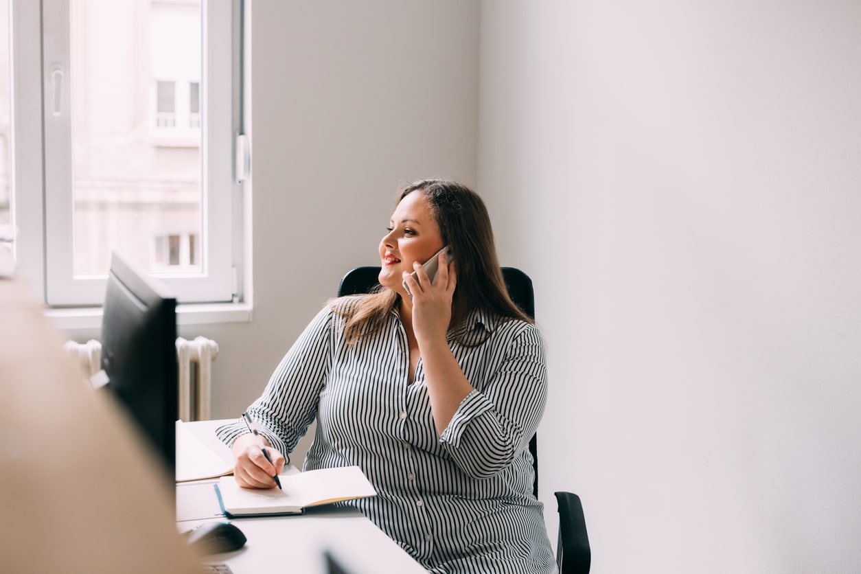 Woman sitting at a desk and talking on the phone. Someone's thumb is in the lens. 