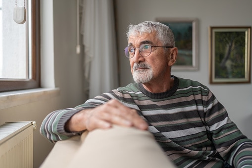 Contemplative older man sitting on his couch at home looking out the window.