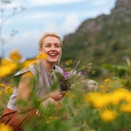 A cheerful woman holding wildflowers sits amidst a beautiful meadow with yellow blossoms and mountains in the background, capturing a sense of peace and connection with nature.