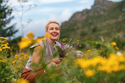 A cheerful woman holding wildflowers sits amidst a beautiful meadow with yellow blossoms and mountains in the background, capturing a sense of peace and connection with nature.